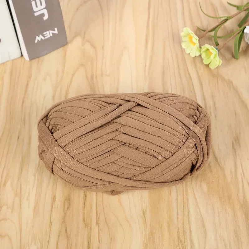 A ball of tan flat thread yarn is lying on a light wooden surface next to some greenery.