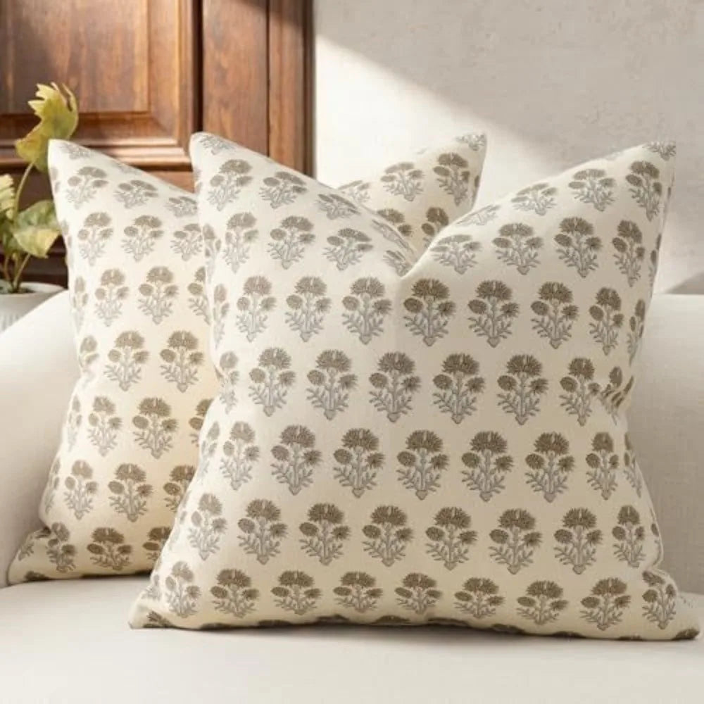 A beige throw pillow cover with a repeating gray floral pattern resting on a white sofa.