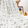 A white muslin swaddle blanket with a green botanical print draped on a surface next to a wooden stool.