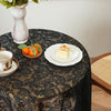 A dark lace tablecloth is set for a meal with an orange fruit arrangement and a slice of cake.