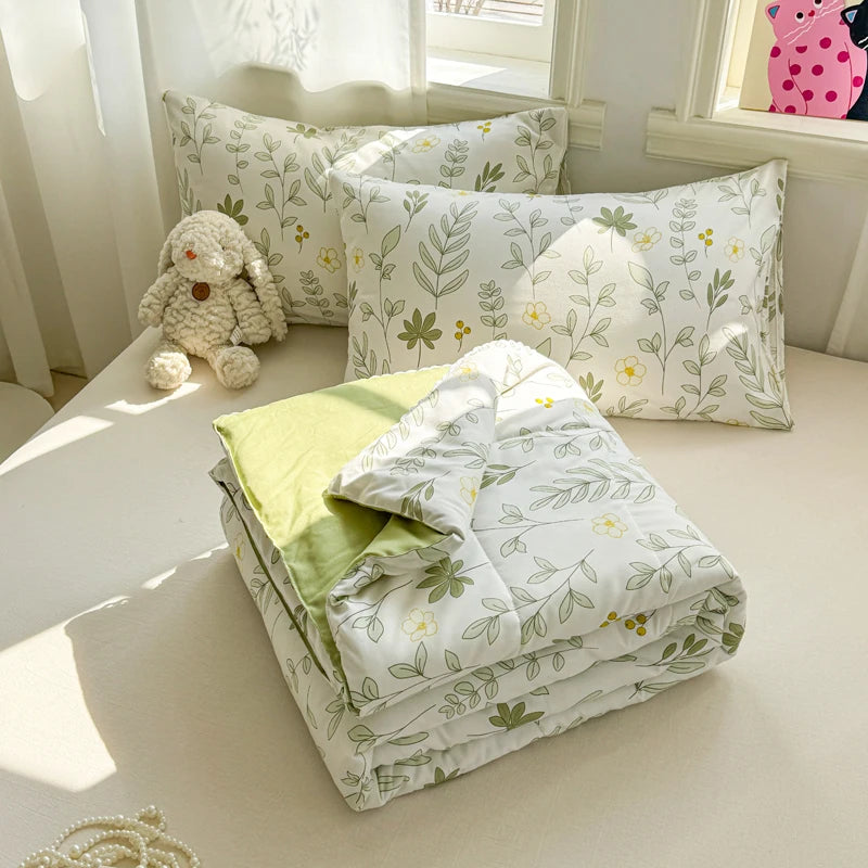 A stack of white and green quilted blankets and pillows with leafy patterns displayed on a light-colored surface.