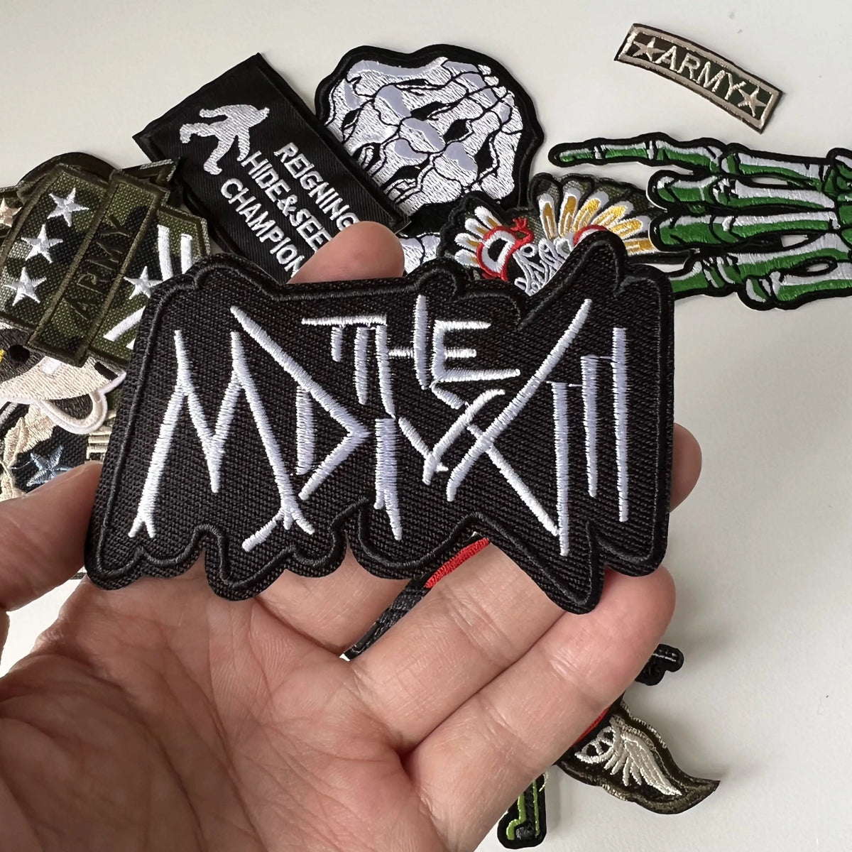 A hand holds several black embroidered patches featuring text and military insignia.