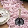 A tall white candle in a black holder rests on a pink crinkle cheesecloth table runner next to silverware.