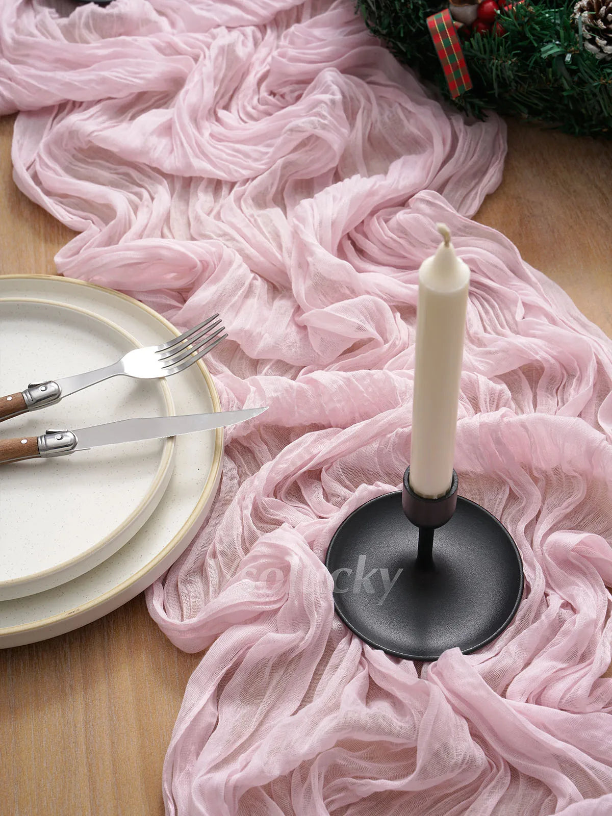 A tall white candle in a black holder rests on a pink crinkle cheesecloth table runner next to silverware.
