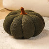 A plush, olive green pumpkin throw pillow with a textured surface rests on a light fabric.