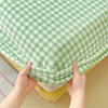 Hands are pulling up a green and white checkered flannel fitted sheet with deep pockets.