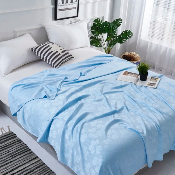 Light blue bamboo fiber cooling blanket draped over a white bed with pillows and greenery.