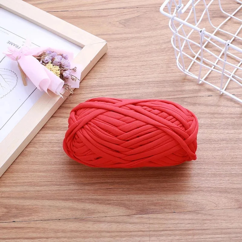 A ball of bright red flat thread yarn lying on a wooden surface next to a white wire mesh basket and a framed flower arrangem
