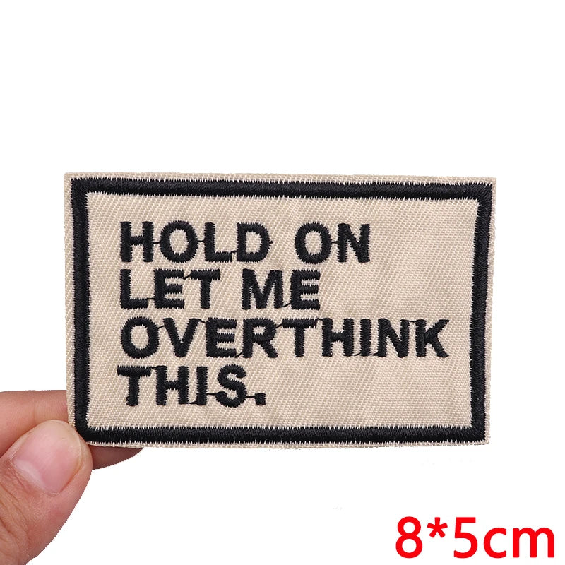 A beige embroidered patch with black text that reads Hold on let me overthink this.