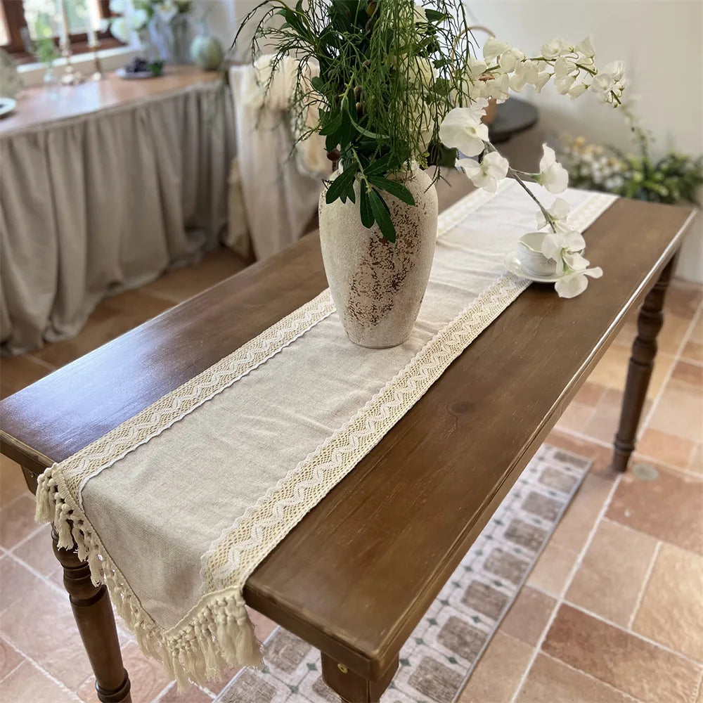 A crochet macrame lace table runner displayed on a wooden table with greenery and white flowers.