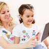 A blonde woman and a young girl smiling while looking at a tablet displaying assorted cartoon embroidery patches.
