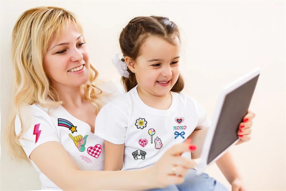 A blonde woman and a young girl smiling while looking at a tablet displaying assorted cartoon embroidery patches.