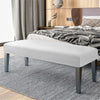 A modern white bench with dark legs and a slipcover is placed on a gray striped rug in a bedroom setting.
