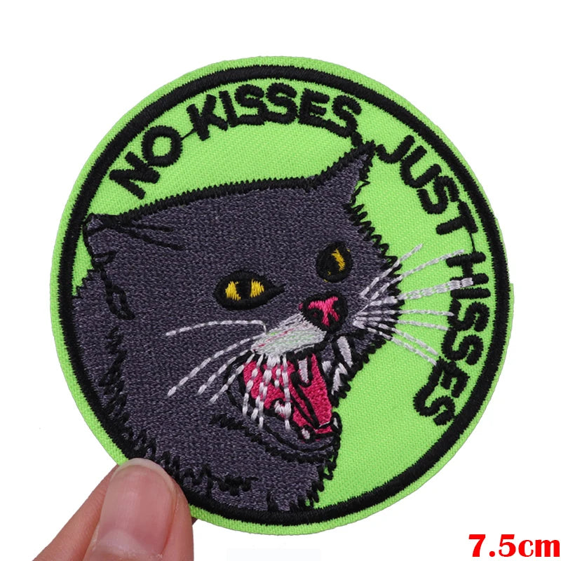 A round embroidered patch featuring a black cat with yellow eyes and white whiskers, surrounded by the text NO KISSES JUST HI