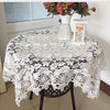 White floral lace tablecloth covering a round table with a vase of flowers and a candle holder.