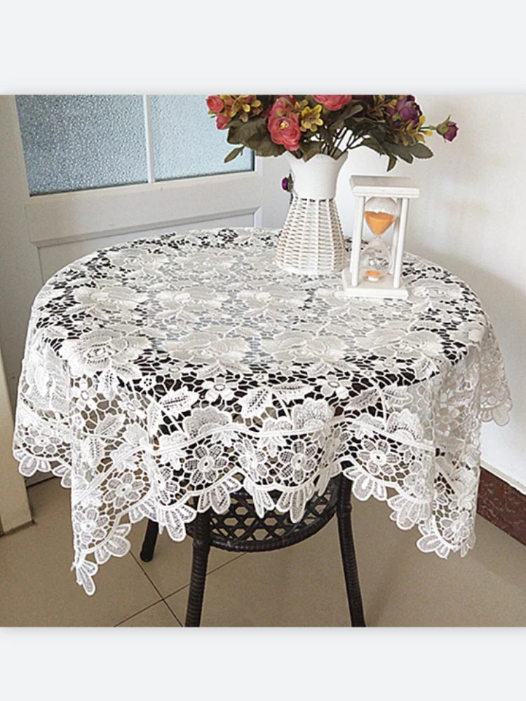 White floral lace tablecloth covering a round table with a vase of flowers and a candle holder.