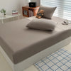 A fitted bed sheet cover in a light gray color is placed on a bed, with pillows and books visible nearby.