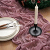 A rustic holiday setting featuring a candle and festive greenery on a pink crinkled fabric table runner.