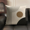 A beige cushion cover featuring black geometric lines and a circular tan accent on a dark sofa.
