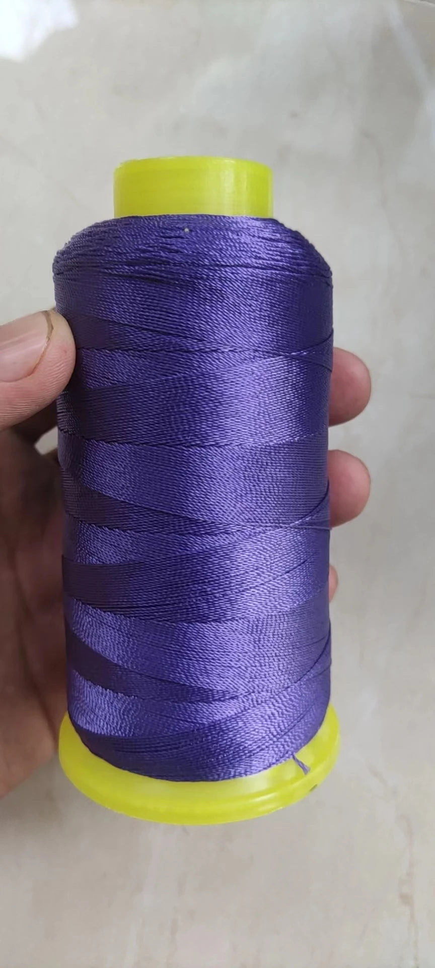 A spool of purple high tenacity polyester sewing thread with a yellow plastic core is held in a hand.