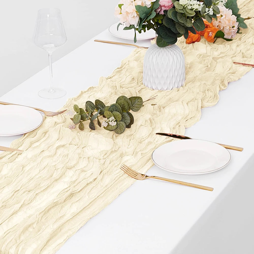 A white table setting featuring a beige boho gauze table runner, plates, silverware, and floral arrangement.