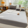 A light gray fitted bed sheet is laid out on a bed with pillows and a stuffed animal in front of a window.