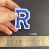 A hand holds a blue and white embroidered letter R patch next to a ruler showing its dimensions.