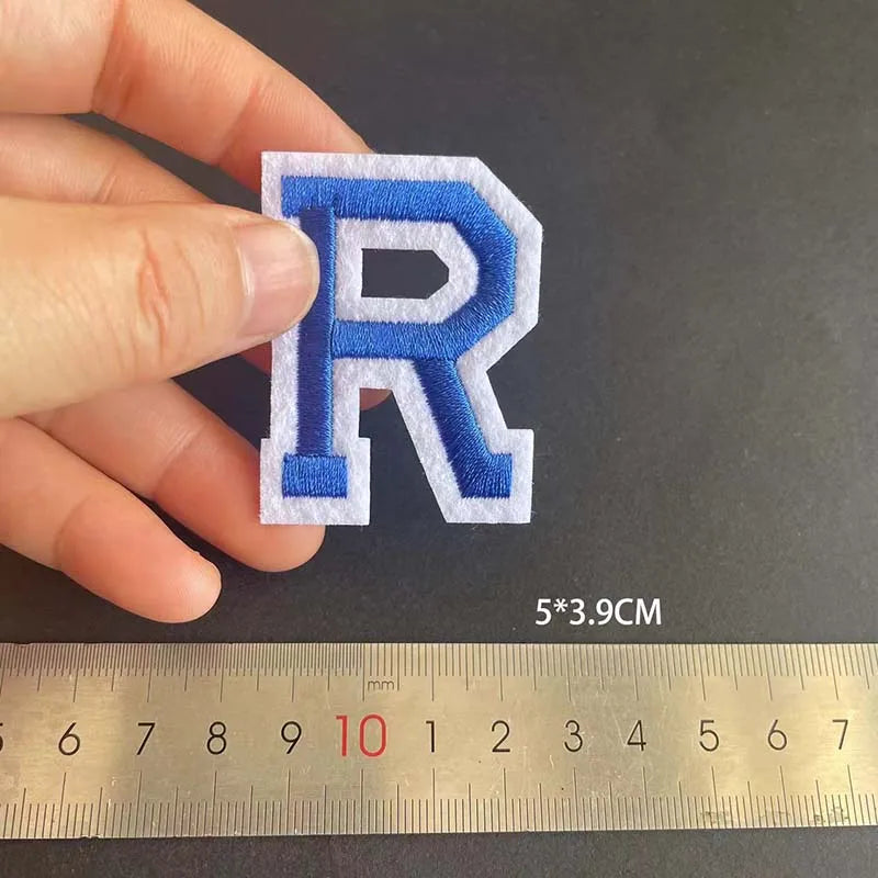 A hand holds a blue and white embroidered letter R patch next to a ruler showing its dimensions.