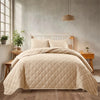 A beige quilted bedspread set is displayed on a bed in a bright room with natural light and wooden furniture.