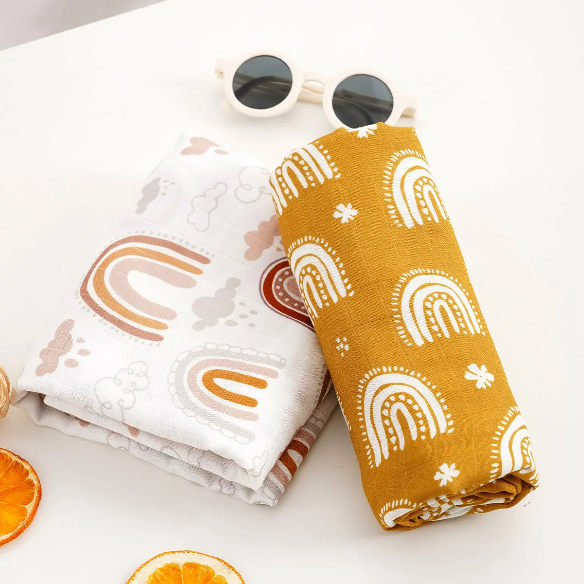 A yellow muslin swaddle blanket with a rainbow pattern is displayed next to sunglasses and an orange slice.