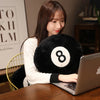 A young woman is using a laptop while resting her chin on a black 8 ball plush pillow.