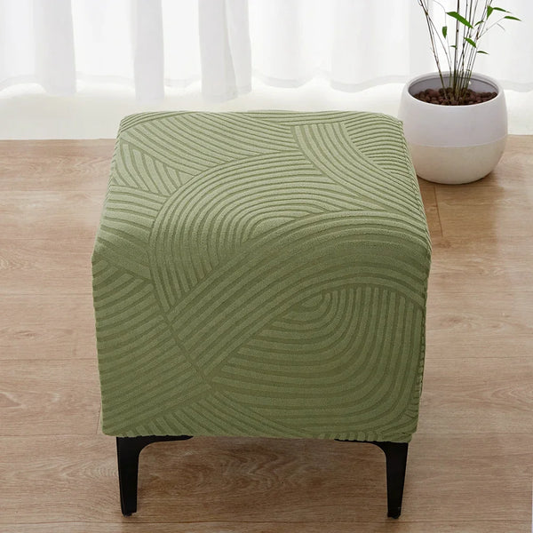 A square ottoman slipcover in olive green with a geometric, wavy pattern resting on a wooden floor.