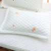 A white quilted pillow with small peach-colored embroidered shapes rests on a white textured surface.