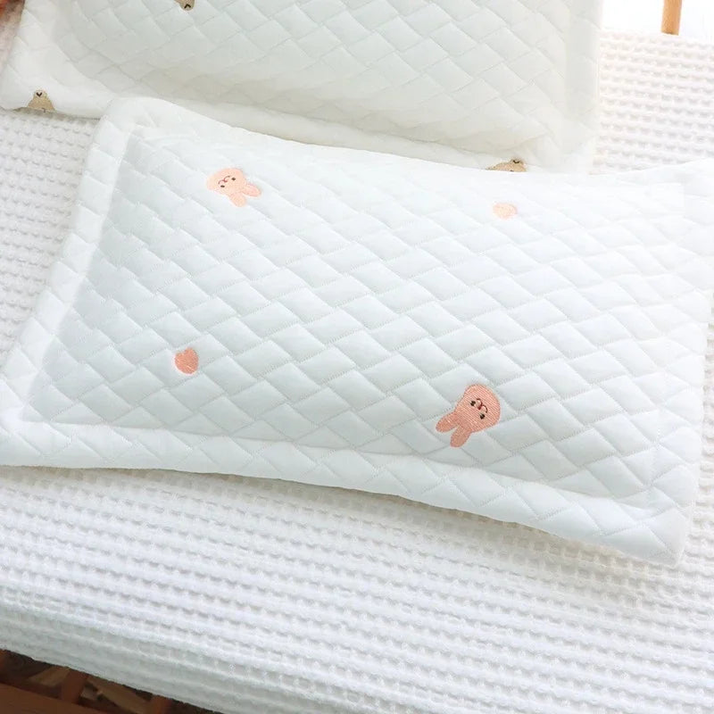 A white quilted pillow with small peach-colored embroidered shapes rests on a white textured surface.