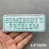 Light mint green embroidered patch with the text Somebody's Problem on a gray fabric background