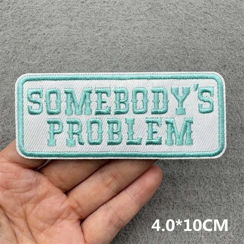 Light mint green embroidered patch with the text Somebody's Problem on a gray fabric background