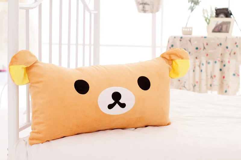 An orange Rilakkuma plush bed pillow with black eyes and yellow ears resting on a white bed.