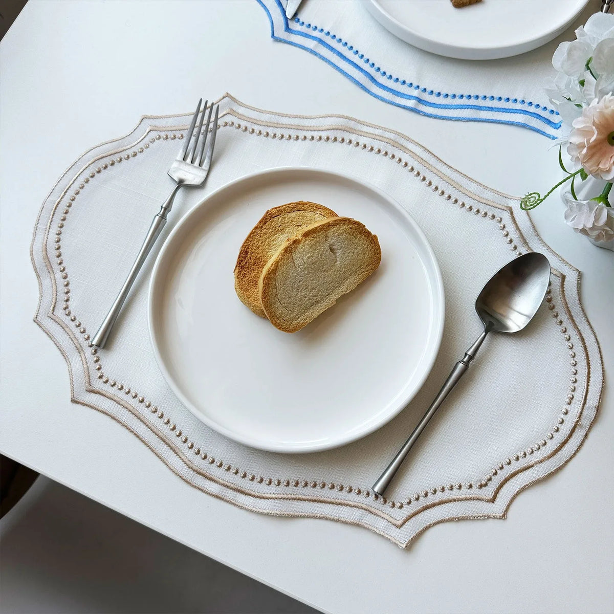 A white plate with a slice of bread, a fork, and a spoon set on an embroidered linen placemat.