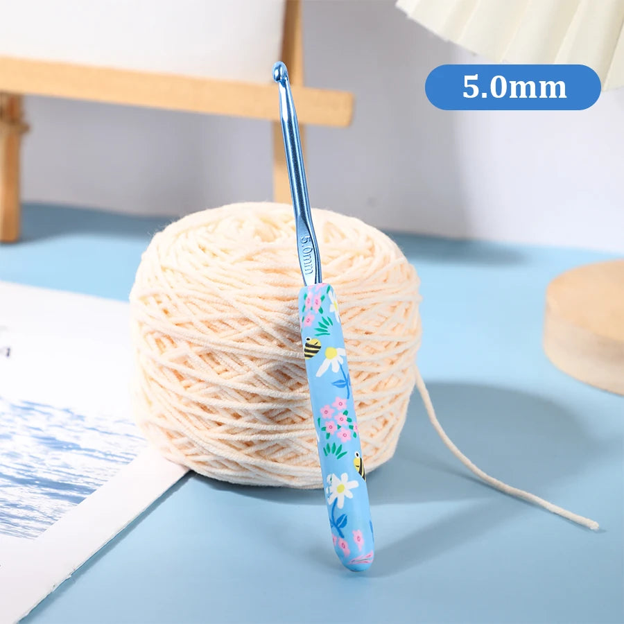 A blue ergonomic crochet hook with a floral design is shown next to a ball of light-colored yarn.