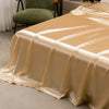 A luxurious satin bedding set in beige color draped over a bed, with a green upholstered chair visible in the background.