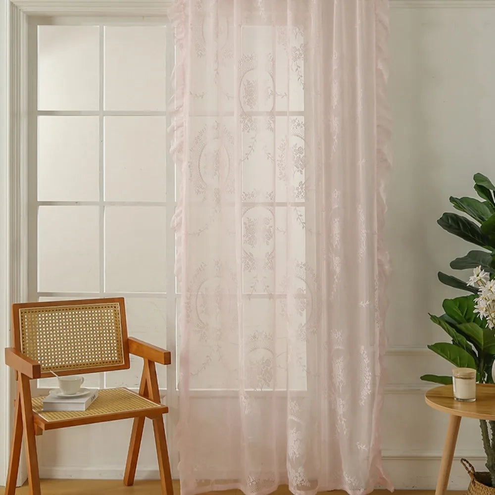 A sheer pink floral lace curtain hangs in a white-framed window next to a wooden chair.