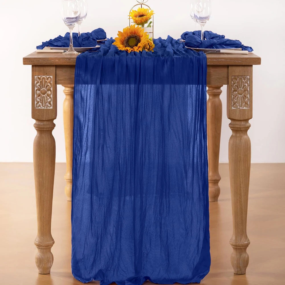 A deep blue lilac cheesecloth table runner draped over a wooden table with sunflowers and glassware.