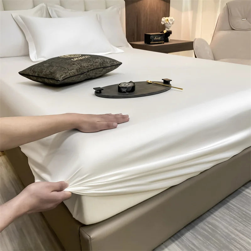 A hand is adjusting a white fitted bed sheet on a bed with a dark pillow and accessories.