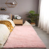 A pink fluffy rug is placed on a light gray floor next to a bed with white linens and wooden decor.
