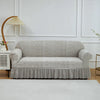 A light gray jacquard stretch sofa cover displayed on a white sofa against a white wall with a wooden lamp.