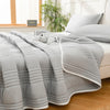 A light gray quilted blanket is draped over a bed, with a green plant visible in the background.