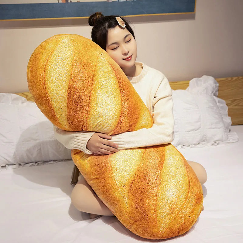 A woman is holding a large, golden-brown soft plush French baguette pillow on her lap in a bright bedroom setting.