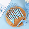 A wooden board holds a set of light blue and white ergonomic crochet hooks for knitting and weaving