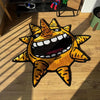 A yellow and orange sun motif floor mat with a fierce expression is placed on a wooden floor.