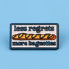 An enamel pin featuring the text less regrets more baguettes on a rectangular background.